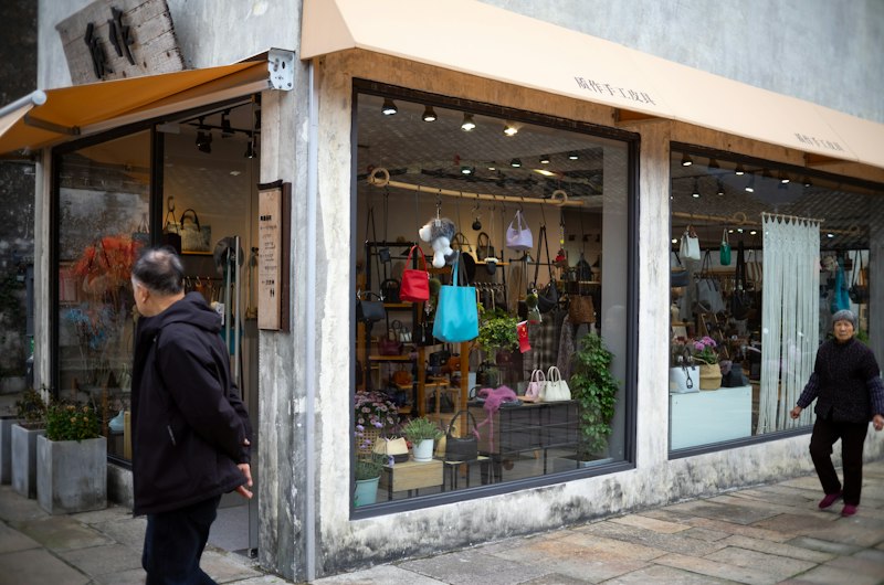 Color photograph of a street-facing fashion and accessories storefront, with handbags, clothing and other merchandise displayed in large windows; two pedestrians are outside. The shop is a small-to-medium street-level retail venue, shown in soft daylight with some warm interior and awning tones.